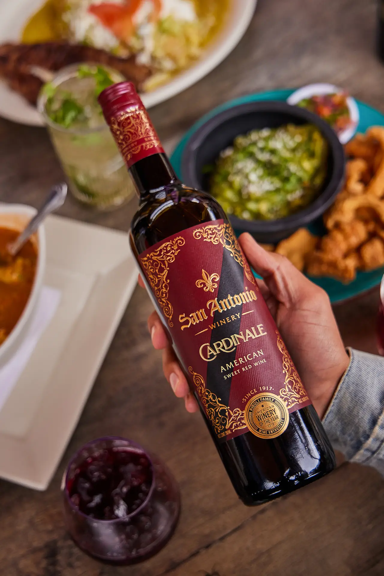 Hand holding a bottle of San Antonio Winery Cardinale over a table set with various dishes.