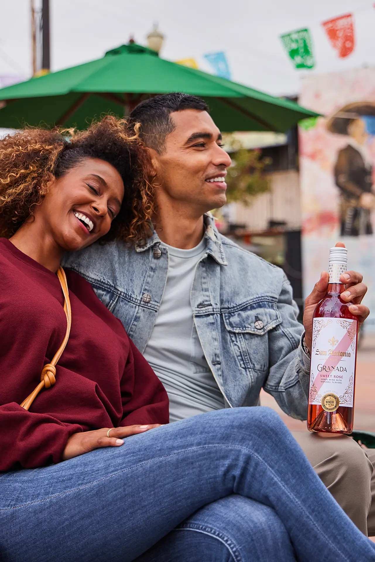 A couple sitting together outdoors, smiling and holding a bottle of San Antonio Winery Granada Sweet Rosé.