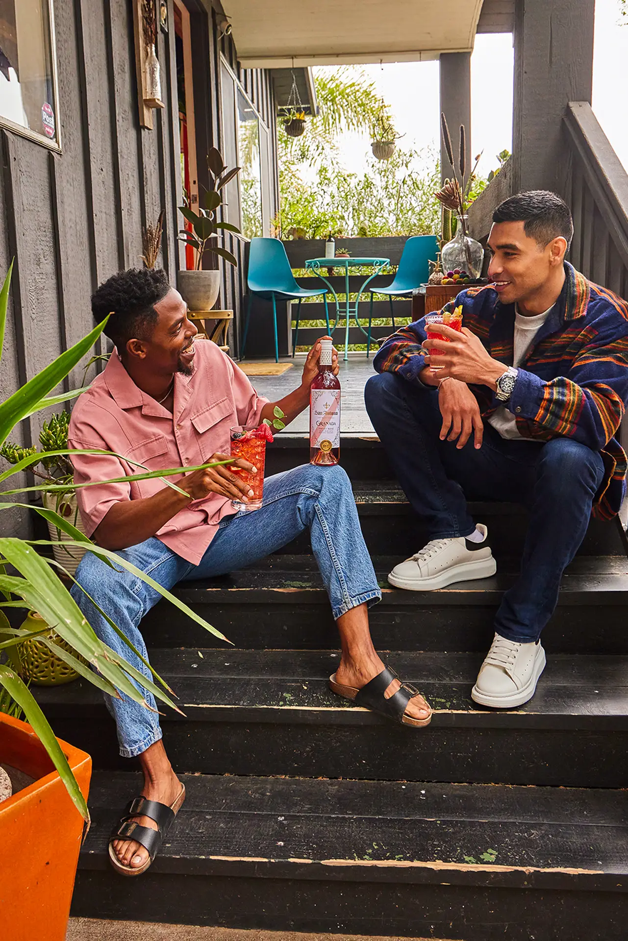 Two friends sitting on porch steps, each holding a glass of red drink, with a bottle of San Antonio Winery wine on the step between them.