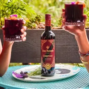 Two people holding up glasses of red wine over a bottle of San Antonio Winery Cardinale on a small, colorful outdoor table.