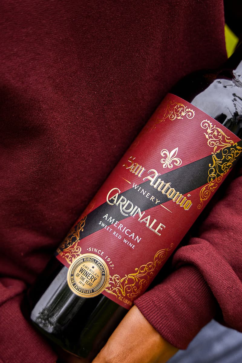 A close-up shot of a person holding a bottle of San Antonio Winery Cardinale American Sweet Red Wine against a background of a maroon sweater.