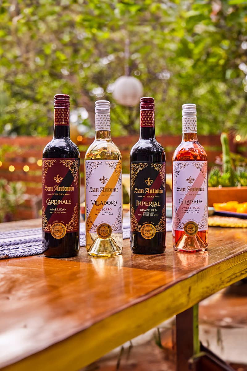 Four bottles of San Antonio Winery wine lined up on a wooden table outdoors. The bottles include Cardinale American Sweet Red Wine, Villadoro Moscato, Imperial Grape Red, and Granada Sweet Rosé.