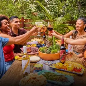 Group of friends raising their glasses for a toast at an outdoor dinner table, with bottles of San Antonio Winery wine and a variety of dishes.