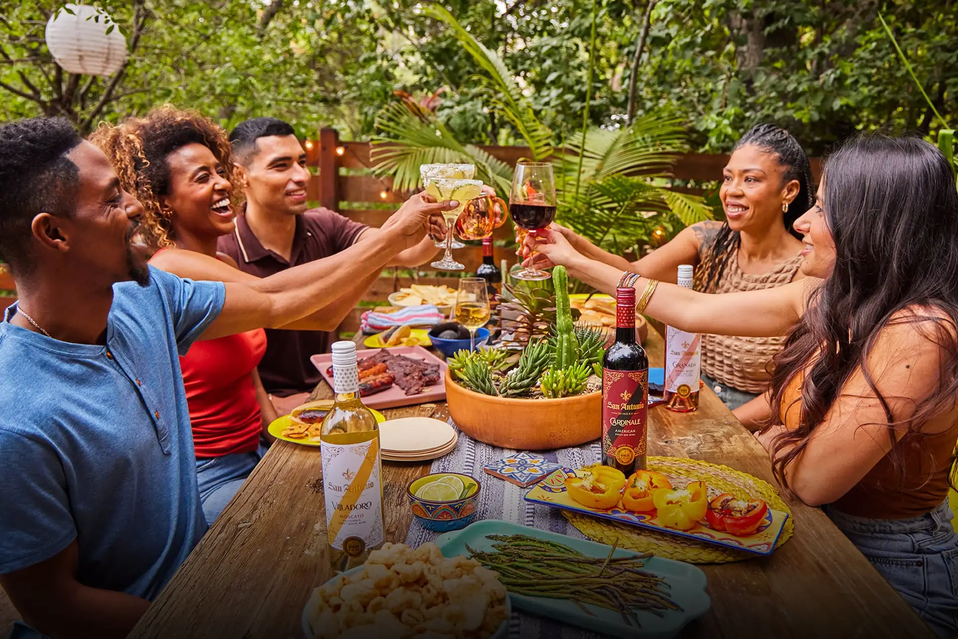 Group of friends raising their glasses for a toast at an outdoor dinner table, with bottles of San Antonio Winery wine and a variety of dishes.