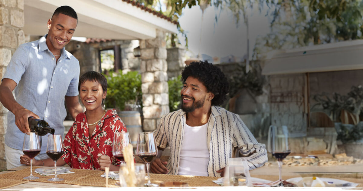Three friends enjoying wine at an outdoor table, with one person pouring wine from a bottle of San Antonio Winery Port California sweet wine.